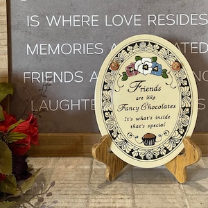 May include: A decorative oval plaque with a floral design and the text "Friends are like Fancy Chocolates. It's what's inside that's special." The plaque is on a wooden stand and has a brown background with black and white accents.