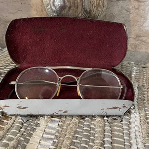 May include: A pair of vintage round glasses with metal frames and clear lenses in a gray metal case with a burgundy velvet lining.