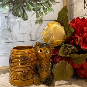 May include: A brown ceramic planter shaped like a beehive with a small brown bear figurine sitting next to it. The planter has a textured surface and a small opening at the bottom. The bear has a brown and black fur pattern and a small pink nose.