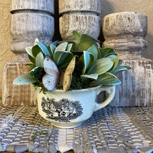 May include: A white teacup with a black and white floral design is filled with green plants and two white butterflies. The teacup is sitting on a white and gray woven rug.
