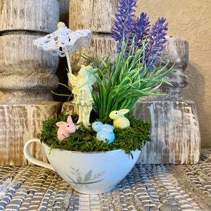 May include: A white teacup filled with moss, a miniature fairy figurine, a small white umbrella, and three pastel colored bunny figurines. The teacup is sitting on a woven rug with a bouquet of lavender flowers in the background.