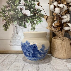 May include: A white ceramic jar with a blue and white scene of a farmhouse and trees. The jar has a round body and a wide mouth.