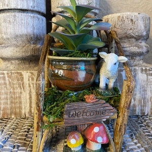May include: A miniature wooden bench with a welcome sign, a small potted succulent plant, a white lamb figurine, and two mushroom figurines. The bench is decorated with moss and a small bird figurine.