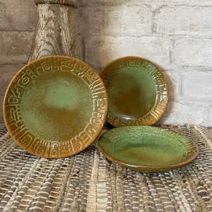 May include: Three green and brown ceramic plates with a geometric design. The plates are on a woven rug.