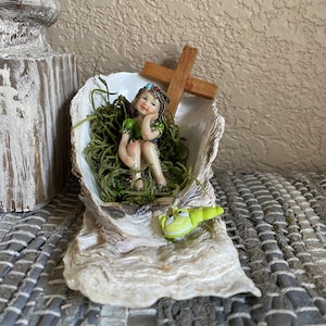 May include: A small fairy figurine sitting in a white seashell with green moss and a wooden cross. A green frog figurine is sitting on the edge of the seashell.