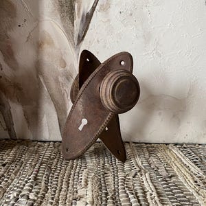 May include: Antique bronze door knobs with keyhole detail. The knobs have a round shape and are attached to oval backplates with decorative edges. The hardware has a vintage, rustic appearance.