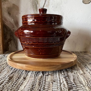 May include: A brown ceramic crock with a lid, featuring a decorative floral design. The crock is sitting on a wooden round serving tray.