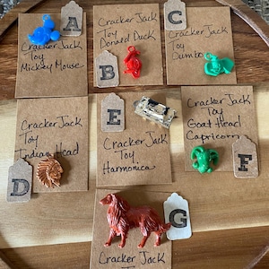 May include: A collection of vintage Cracker Jack toy prizes, including a blue Mickey Mouse, a red Donald Duck, a green Dumbo, a brown Indian Head, a silver harmonica, a green Capricorn, and a brown Lassie dog. Each toy is displayed on a brown card with the Cracker Jack logo and the name of the toy.