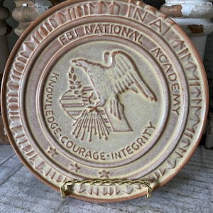 May include: A tan ceramic plate with a brown border and a yellow center. The center features a seal with an eagle, a torch, and the words "FBI National Academy" and "Knowledge, Courage, Integrity".