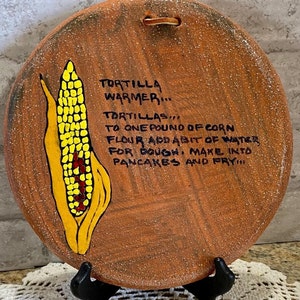 May include: A round, terracotta tortilla warmer with a painted corn cob design. The warmer has a handwritten recipe for making tortillas: "TORTILLA WARMER... TORTILLAS... TO ONE POUND OF CORN FLOUR ADD A BIT OF WATER FOR DOUGH. MAKE INTO PANCAKES AND FRY!"