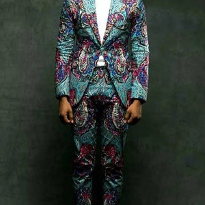 K&ouml;nnte beinhalten: Ein Mann in einem gemusterten Anzug in Blaut&ouml;nen, Rosa und Schwarz. Der Anzug hat ein Paisley-Muster. Er tr&auml;gt eine schwarze Sonnenbrille und schwarze Schuhe.