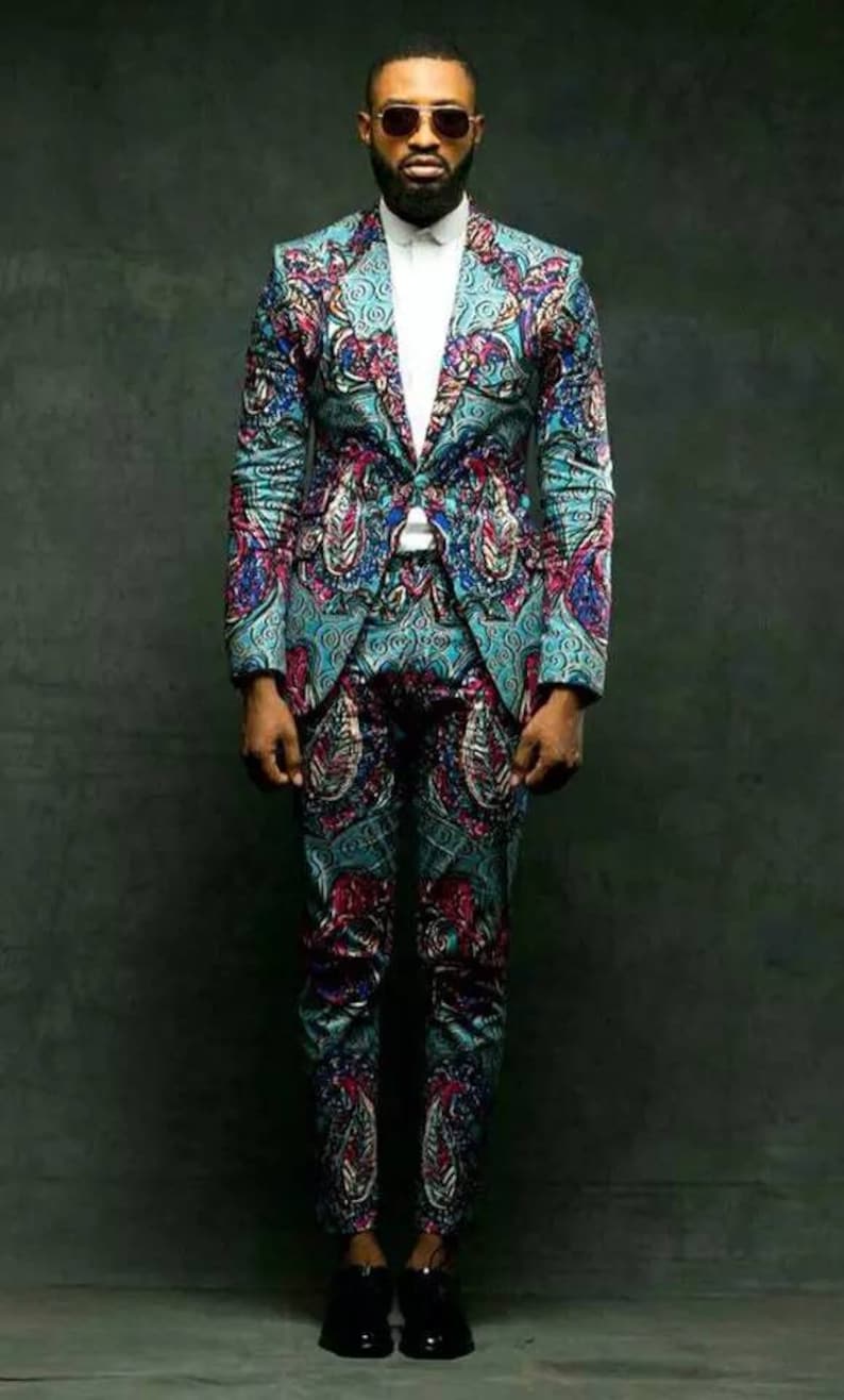 K&ouml;nnte beinhalten: Ein Mann in einem gemusterten Anzug in Blaut&ouml;nen, Rosa und Schwarz. Der Anzug hat ein Paisley-Muster. Er tr&auml;gt eine schwarze Sonnenbrille und schwarze Schuhe.