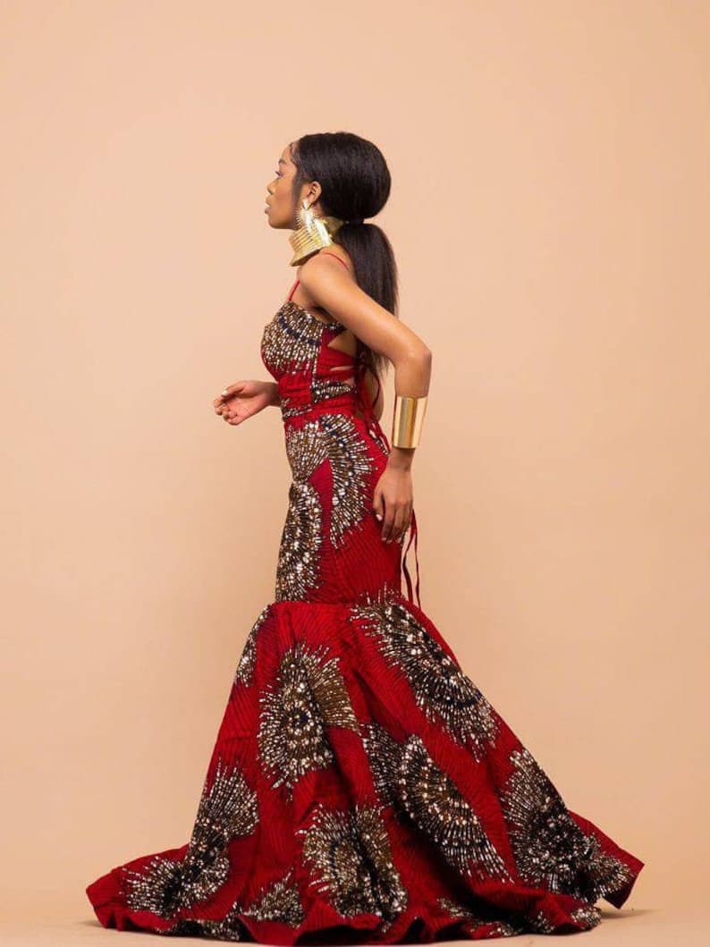 May include: A woman wearing a red, gold, and black patterned dress with a fitted bodice and a flowing skirt. She is wearing gold jewelry and a gold bracelet.
