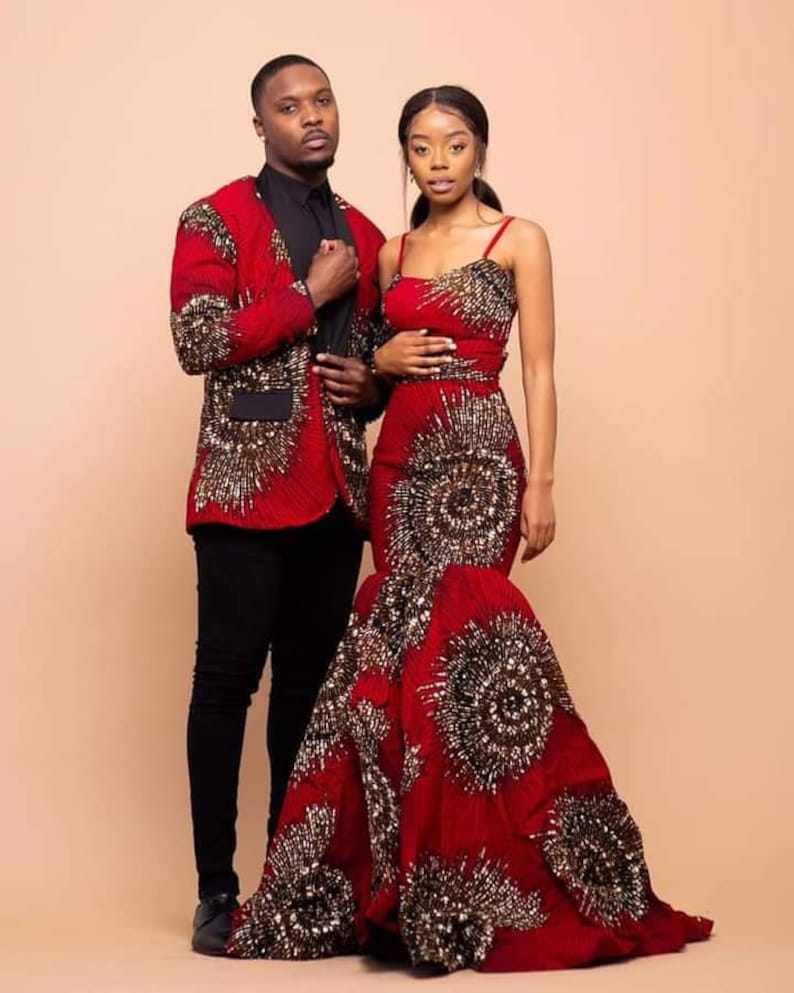 May include: A couple wearing matching red and gold patterned outfits. The woman is wearing a long, fitted dress with a mermaid skirt. The man is wearing a blazer with a black shirt and black pants.