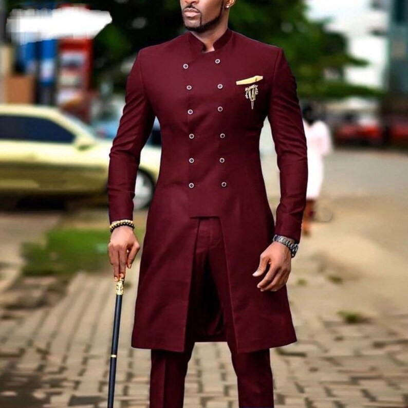 May include: A burgundy suit with a mandarin collar and double-breasted button closure. The long jacket features a split front and a small gold-colored emblem on the chest. The suit is paired with matching trousers.