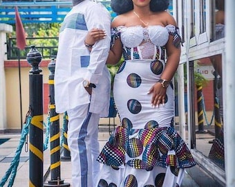 May include: A couple in traditional white clothing with a patterned design. The woman is wearing a white dress with a lace bodice and a colourful patterned skirt. The man is wearing a white shirt with a blue and white patterned design. The couple are standing in front of a building with a blue and white striped awning.