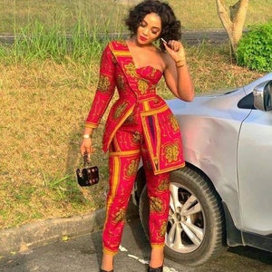 May include: A woman wearing a red and gold patterned pantsuit with a one-shoulder design. She is standing in front of a car and holding a small purse.