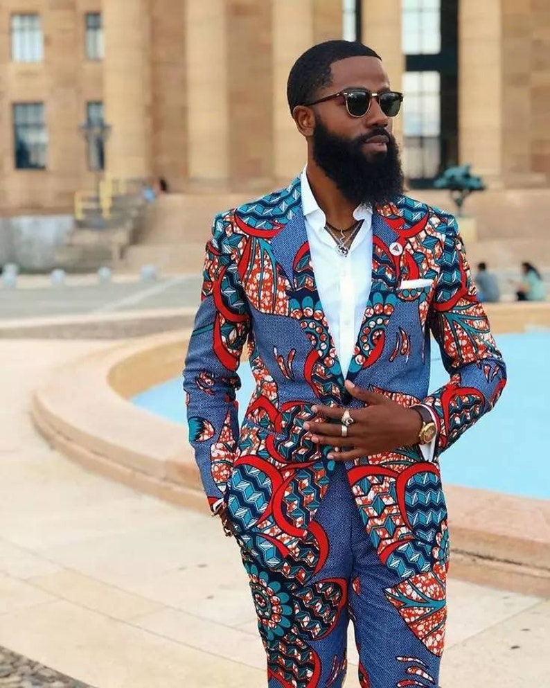 May include: A man wearing a blue, red and white patterned suit jacket and matching trousers. He is wearing a white shirt, sunglasses and a silver necklace.