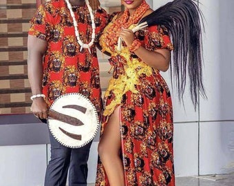 Puede incluir: Una pareja con atuendo tradicional nigeriano. La mujer lleva un vestido rojo y dorado con una abertura alta y un collar de cuentas. El hombre lleva una camisa roja y negra con un collar de cuentas blancas y pantalones negros. Ambos sonríen y sostienen un abanico.