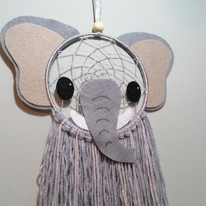 May include: A grey and beige felt elephant dreamcatcher with black eyes and white tusks. The dreamcatcher has a silver hoop and grey and white yarn fringe.