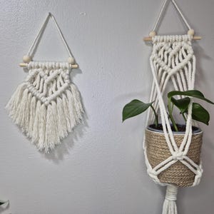 May include: Two macrame plant hangers, one small and one large, with wooden dowels and white cotton cord. The larger hanger is holding a potted plant with green leaves.