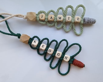 May include: Two personalized Christmas tree ornaments with the names JAMIE and KEOGH spelled out with wooden letter beads. The ornaments are made with green and white string and have wooden beads at the top.
