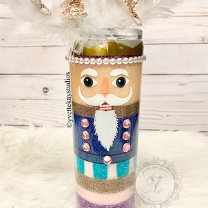 以下が含まれることがあります： ナッツクラッカーのデザインが施されたキラキラ輝くタンブラー。タンブラーは、ゴールドのグリッター、ホワイトのグリッター、ピンクのグリッター、ブルーのグリッター、ターコイズのグリッターで飾られています。タンブラーの上には白い羽根とゴールドのストローが付いています。