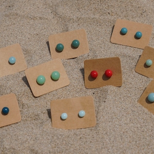 Op de afbeelding: Assortiment van kleine ronde oorbel studs gepresenteerd op bruine kaartjes. De oorbel studs zijn in verschillende tinten blauw, groen en rood.