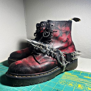 Puede incluir: Botas de cuero negras y rojas con una cadena de púas. Las botas tienen suela de plataforma, cordones negros y un patrón rojo y negro. La cadena de púas rodea el tobillo.