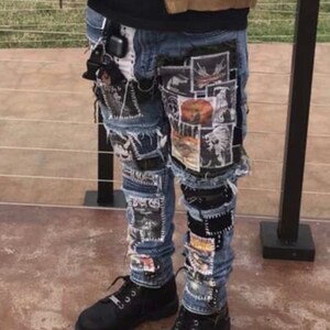 Puede incluir: Vaqueros azules con m&uacute;ltiples parches gr&aacute;ficos y detalles desgastados. Los vaqueros se combinan con zapatos negros. La persona lleva una chaqueta marr&oacute;n y una camiseta negra.