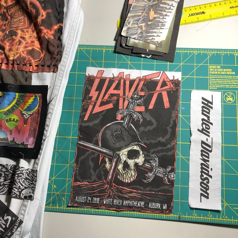 Puede incluir: Un cartel de concierto de Slayer en negro y rojo con una calavera que lleva un casco y espadas cruzadas. El cartel incluye la fecha 24 de agosto de 2018 y el lugar White River Amphitheatre, Auburn, WA. Tambi&eacute;n se ven parches con texto y gr&aacute;ficos.