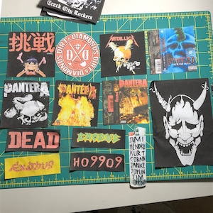 Op de afbeelding: Een verzameling zwarte stoffen patches met bandlogo's en albumhoezen, waaronder Metallica en Pantera. Een aansteker met namen van muzikanten is ook aanwezig. De patches liggen op een groene snijmat.