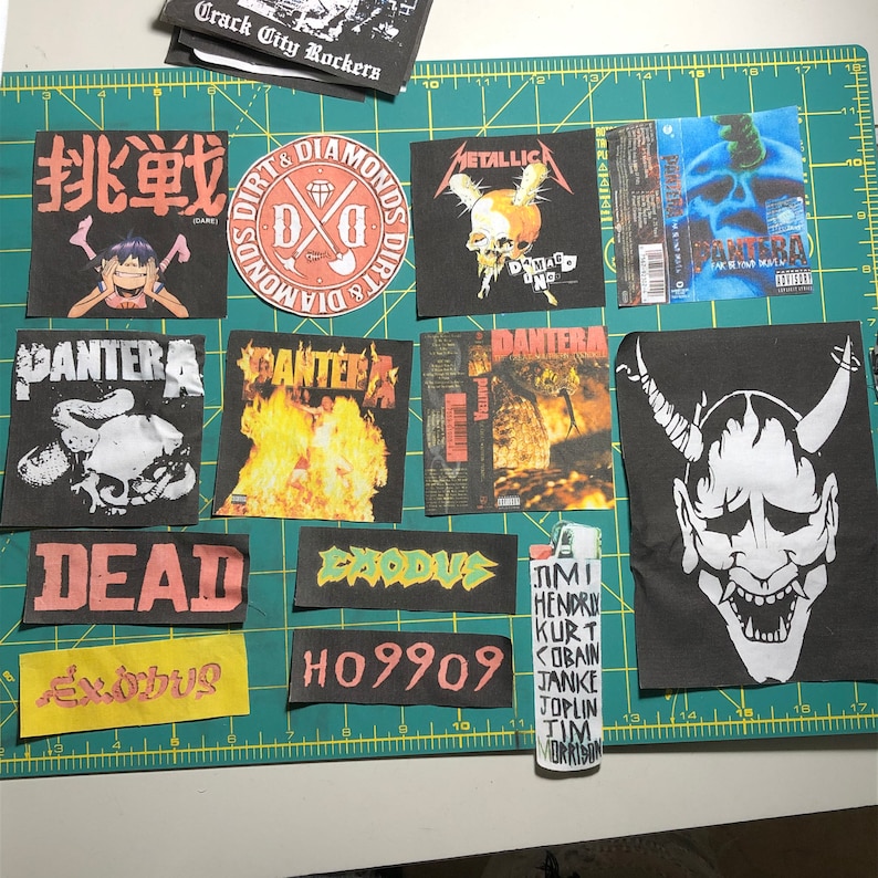 Puede incluir: Un conjunto de parches de tela negra con logotipos de bandas y portadas de &aacute;lbumes, incluyendo Metallica y Pantera. Tambi&eacute;n hay un encendedor con nombres de m&uacute;sicos. Los parches est&aacute;n sobre una esterilla de corte verde.