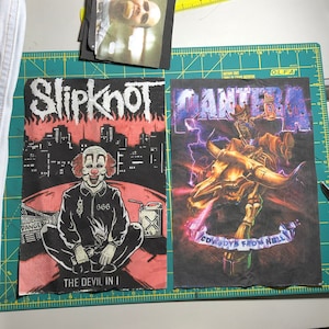 Puede incluir: Dos parches de tela negra con logotipos de bandas. Un parche muestra "Slipknot" sobre una ilustraci&oacute;n de payaso con el texto "THE DEVIL IN I". El otro parche muestra "PANTERA" con un esqueleto montando un toro y el texto "COWBOYS FROM HELL".