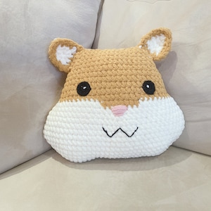 May include: A crocheted hamster pillow with brown fur, white cheeks, and black eyes. The pillow is sitting on a light brown couch.