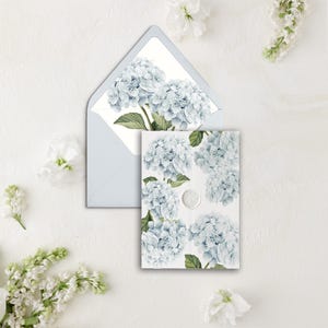 Puede incluir: Un sobre blanco con un diseño floral de hortensias azules en la parte delantera. El sobre está abierto y se puede ver una tarjeta más pequeña con un diseño floral similar en el interior.