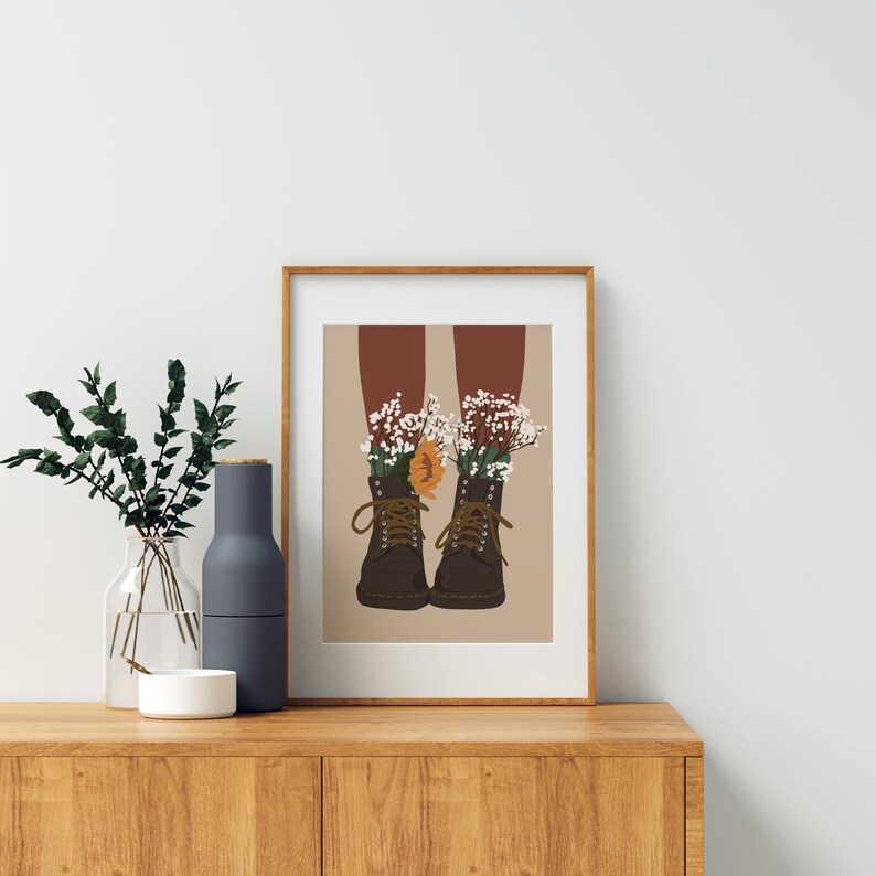 May include: A framed illustration of a pair of brown boots with white and orange flowers growing out of them.