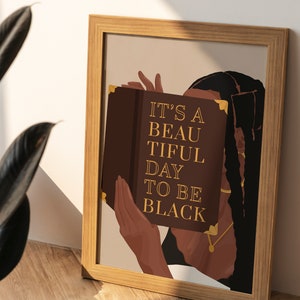 May include: A framed print with a brown background featuring a woman with long black braids holding a book in front of her face. The book has gold accents and the text "IT'S A BEAUTIFUL DAY TO BE BLACK" in gold lettering.