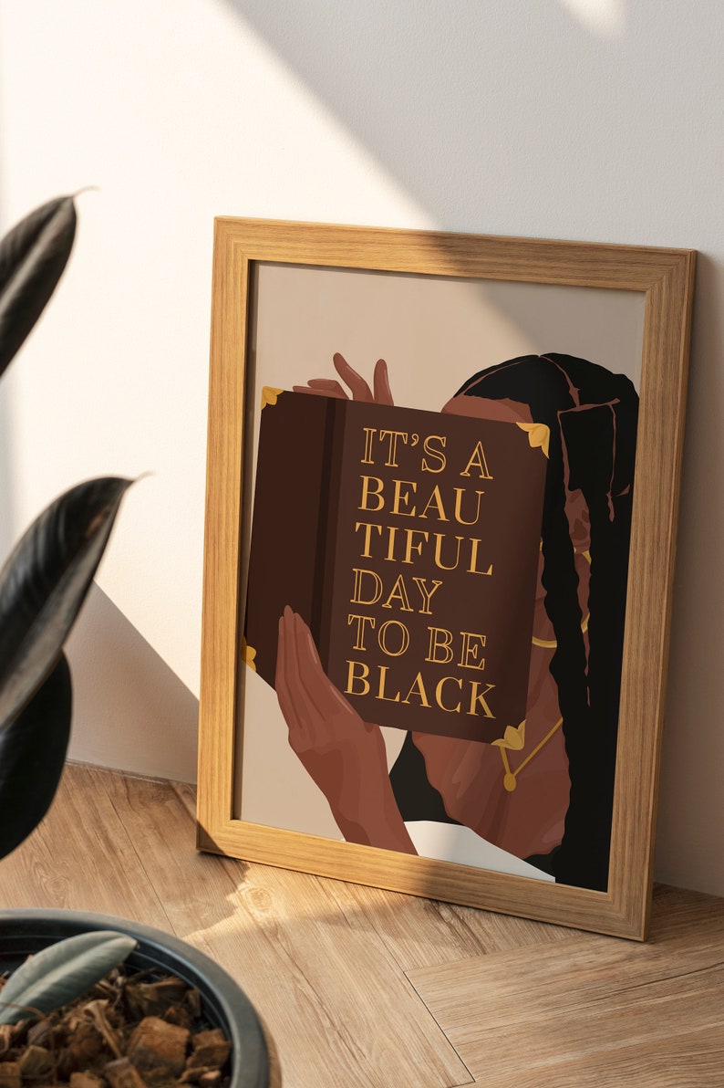 May include: A framed print with a brown background featuring a woman with long black braids holding a book in front of her face. The book has gold accents and the text "IT'S A BEAUTIFUL DAY TO BE BLACK" in gold lettering.