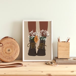 May include: A framed illustration of a pair of brown boots with white and orange flowers growing out of them. The boots are on a beige background.