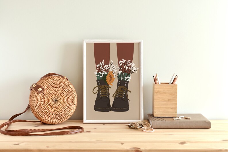 May include: A framed illustration of a pair of brown boots with white and orange flowers growing out of them. The boots are on a beige background.