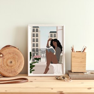 May include: A digital illustration of a Black woman sitting on a balcony, reading a book, with a view of city buildings in the background. The illustration is framed in a white frame.