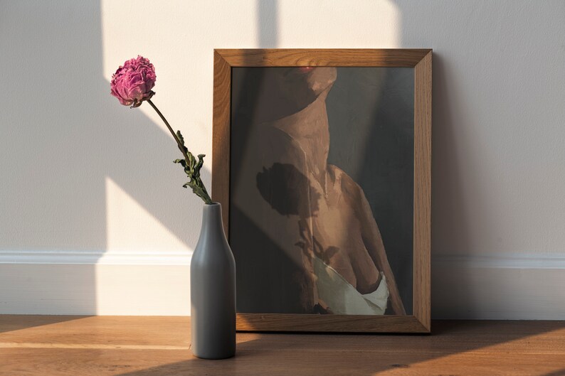 May include: A framed print of a nude figure with a dark background. A single dried pink peony flower in a gray vase sits in front of the print.