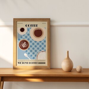 May include: A framed art print featuring coffee cups and the text "COFFEE" and "WE LOVE COFFEE." The artwork has a light blue checkered background and a light brown wooden frame. A wooden vase and ball sit on a wooden table.