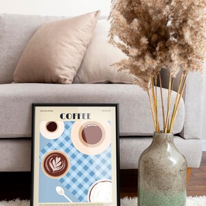 May include: A framed coffee-themed art print with a black border, featuring coffee cups and the text "COFFEE" and "WE LOVE COFFEE". The print is displayed on a white rug, with a gray sofa and decorative vase in the background.