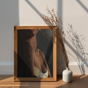 May include: A framed painting of a person's torso with a dark background. The painting is in a wooden frame and is hanging on a white wall. A vase with dried flowers is in the foreground.