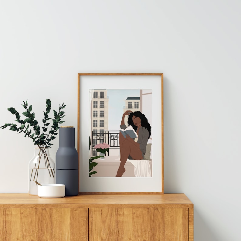 May include: A framed illustration of a woman sitting on a balcony reading a book. The woman has long dark hair and is wearing a light blue shirt. The balcony overlooks a city street with buildings in the background.