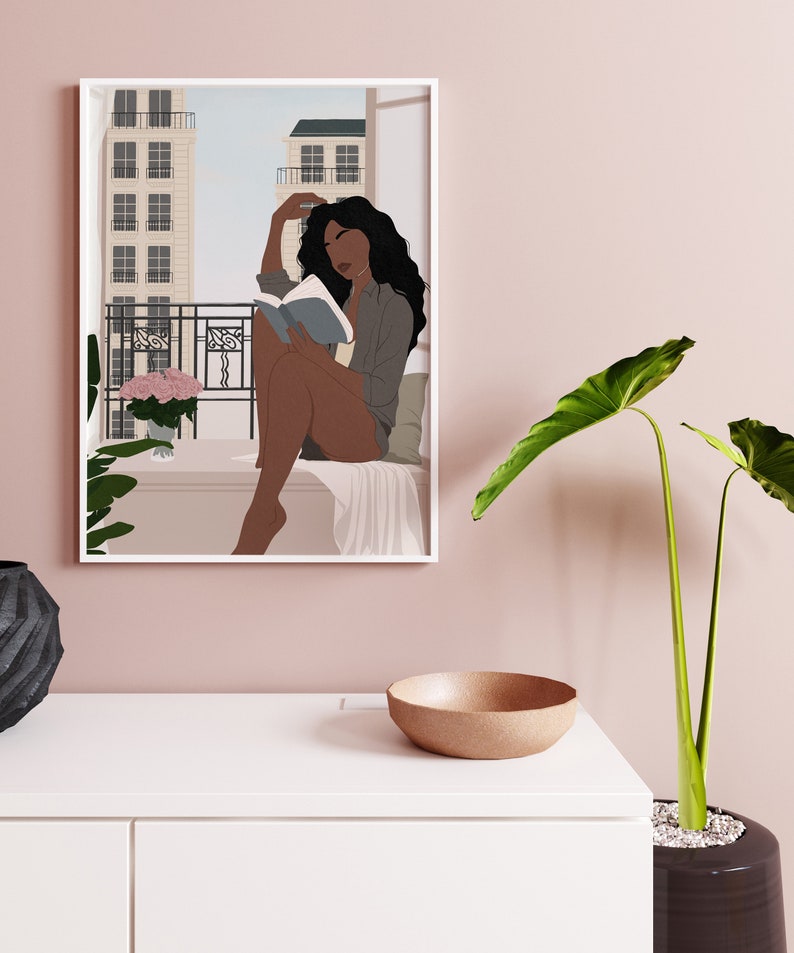 May include: A framed illustration of a woman sitting on a balcony reading a book. The woman has dark skin and long black hair. She is wearing a blue shirt and has a pink flower bouquet on the balcony.