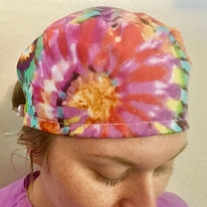 May include: A colorful tie-dye scrub hat with a swirl pattern in shades of pink, orange, yellow, blue, and green. The hat is made of soft, comfortable fabric and is perfect for nurses, doctors, and other healthcare professionals.