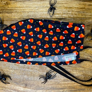 Op de afbeelding: Zwart gezichtsmasker met een snoepmaispatroon en zwarte elastische banden. Het masker heeft een wit label met de tekst "Sewn with Love & Sparkle".